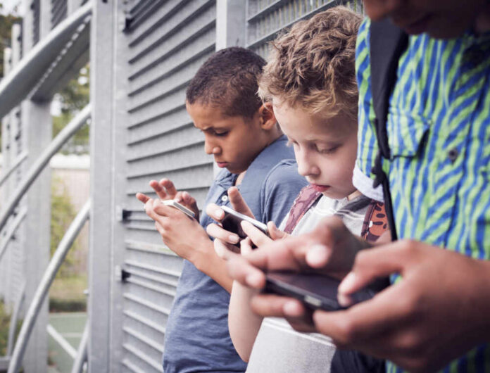 Three children focused on their smartphones while standing outdoors