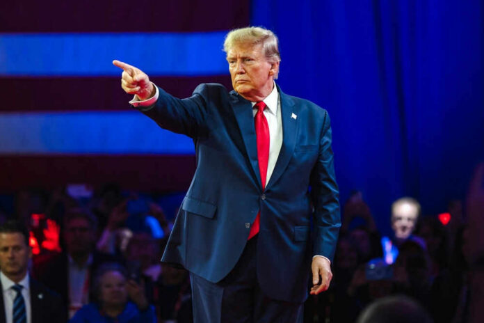 trumpid2430065795 Person in suit pointing on stage with audience watching