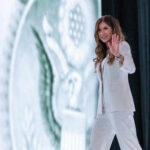 Trump Yanks DHS Chief—What Broke? Person in white suit waves on stage background