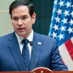 U.S.-Israeli Airpower Surge: What’s Next? A man in a blue suit speaking into a microphone with an American flag in the background