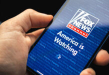LIVE Suplex Shocker: Fox’s Hornacek Smiles Through It Hand holding smartphone showing Fox News Channel logo.
