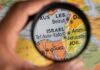 A hand holding a magnifying glass over a map focusing on Israel