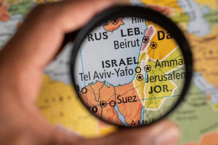 A hand holding a magnifying glass over a map focusing on Israel