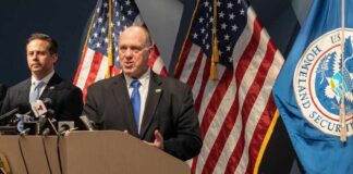 Government officials speaking at a press conference with U.S. flags in the background