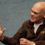 A man in a brown jacket speaking into a microphone with a smile