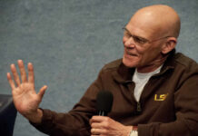 A man in a brown jacket speaking into a microphone with a smile