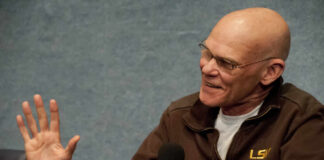 A man in a brown jacket speaking into a microphone with a smile