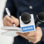 A journalist holding a notebook and microphones labeled 'NEWS'