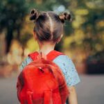 A young child with a red backpack seen from behind, standing outdoors