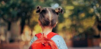 A young child with a red backpack seen from behind, standing outdoors