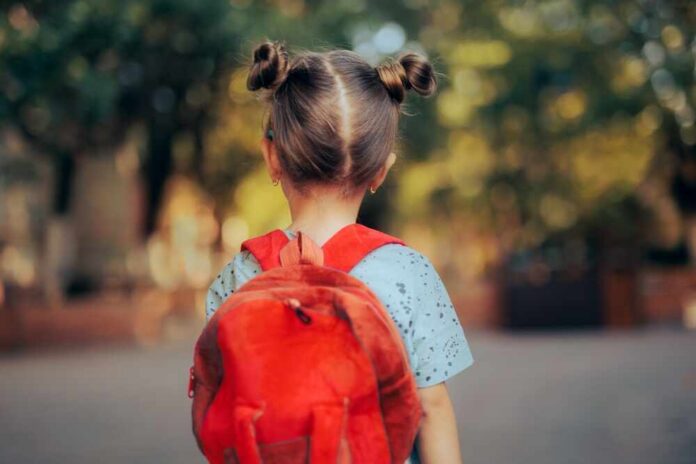 A young child with a red backpack seen from behind, standing outdoors