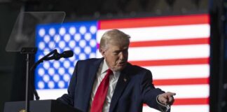 A man in a suit gestures while speaking at a podium with an American flag in the background