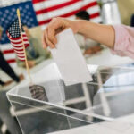 Hand placing ballot in box with American flag nearby