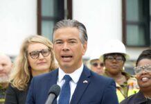 Man in a navy suit speaking at a microphone with people standing behind