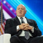 Man in suit sitting on stage with American flag.