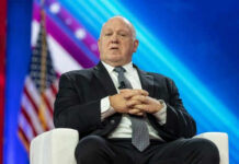Death Threats Explode After ‘Nazi’ Smears Target ICE Man in suit sitting on stage with American flag.