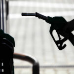 Hormuz Reopens—Markets Panic Anyway Silhouette of a hand holding a fuel nozzle at a gas station