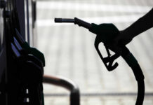 Hormuz Reopens—Markets Panic Anyway Silhouette of a hand holding a fuel nozzle at a gas station