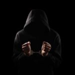A hooded figure with hands in handcuffs against a dark background