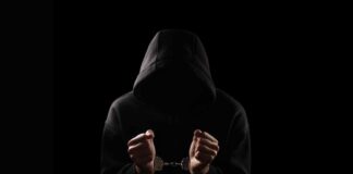 A hooded figure with hands in handcuffs against a dark background