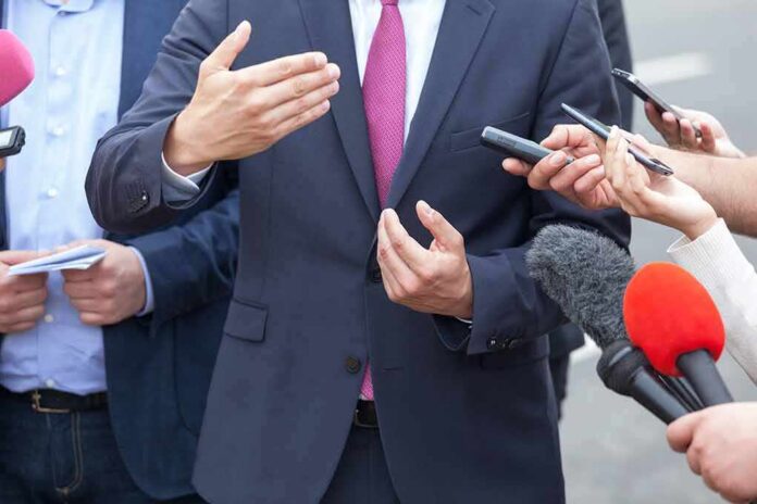 shutterstock_1040735278 Businessperson in suit speaking to reporters with microphones