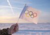 IOC Bends — Russia Sneaks Back A hand holding an Olympic flag against a snowy landscape during sunset