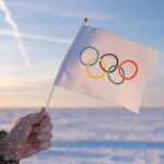 IOC Bends — Russia Sneaks Back A hand holding an Olympic flag against a snowy landscape during sunset