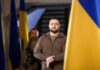 man in olive jacket standing between large blue and yellow flags