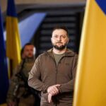 Funeral Becomes High-Stakes Power Play man in olive jacket standing between large blue and yellow flags