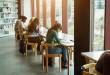 Students studying at long tables by large library windows