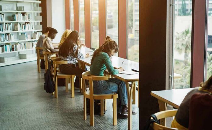 Students studying at long tables by large library windows