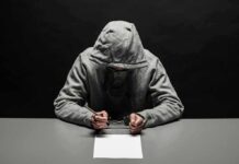 77 Break-Ins—Families On Edge Person in gray hoodie writing while handcuffed at a table