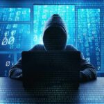 Water Systems Breached — Who’s Behind It? Hooded figure using a laptop in front of glowing code