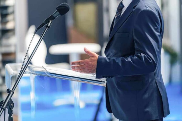 shutterstock_2692761339 Speaker in a suit at a podium with microphone