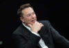 Elon Musk speaking on stage in a dark suit, resting his chin on his hand