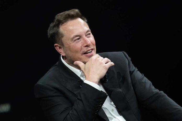 Elon Musk speaking on stage in a dark suit, resting his chin on his hand
