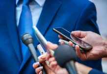Person in a blue suit surrounded by reporters’ microphones and smartphones
