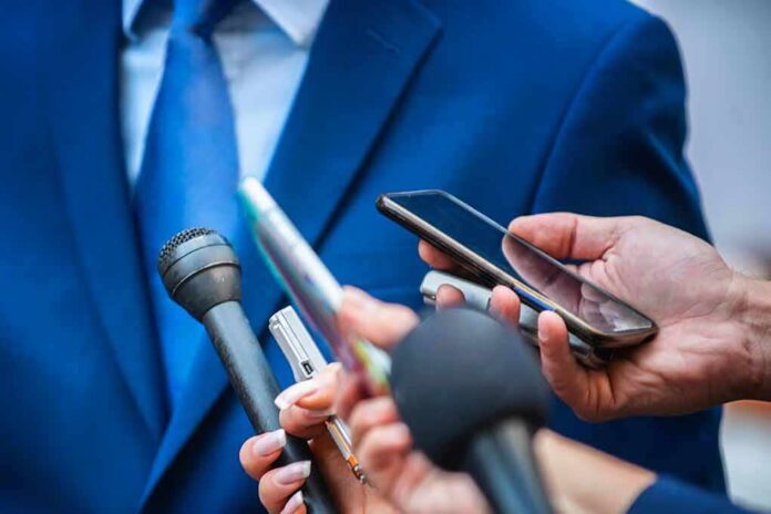 shutterstock_1333000841 Person in a blue suit surrounded by reporters’ microphones and smartphones