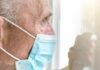 Older man wearing a surgical mask looking out a window