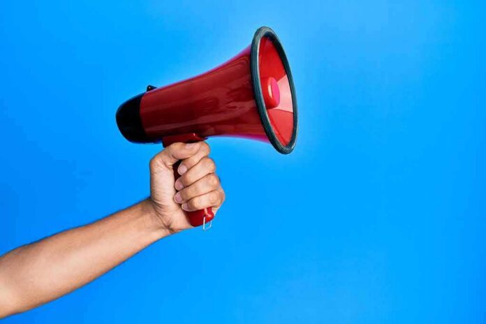 shutterstock_1890023353 Hand holding a red megaphone against a blue background