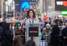 Kathy Hochul speaks at a clear podium in a city street; Josh Boyce stands among the crowd
